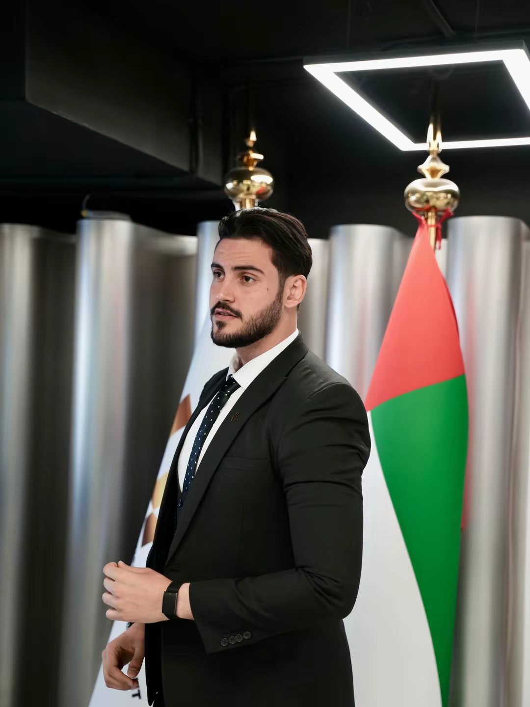 Mahmoud Ashri in a black suit standing before national flags at an official reception